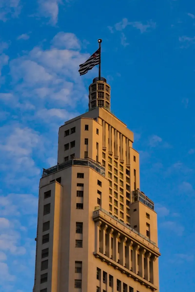 Edifício Altino Arantes ao entardecer, com a bandeira paulista, sob céu azul com nuvens