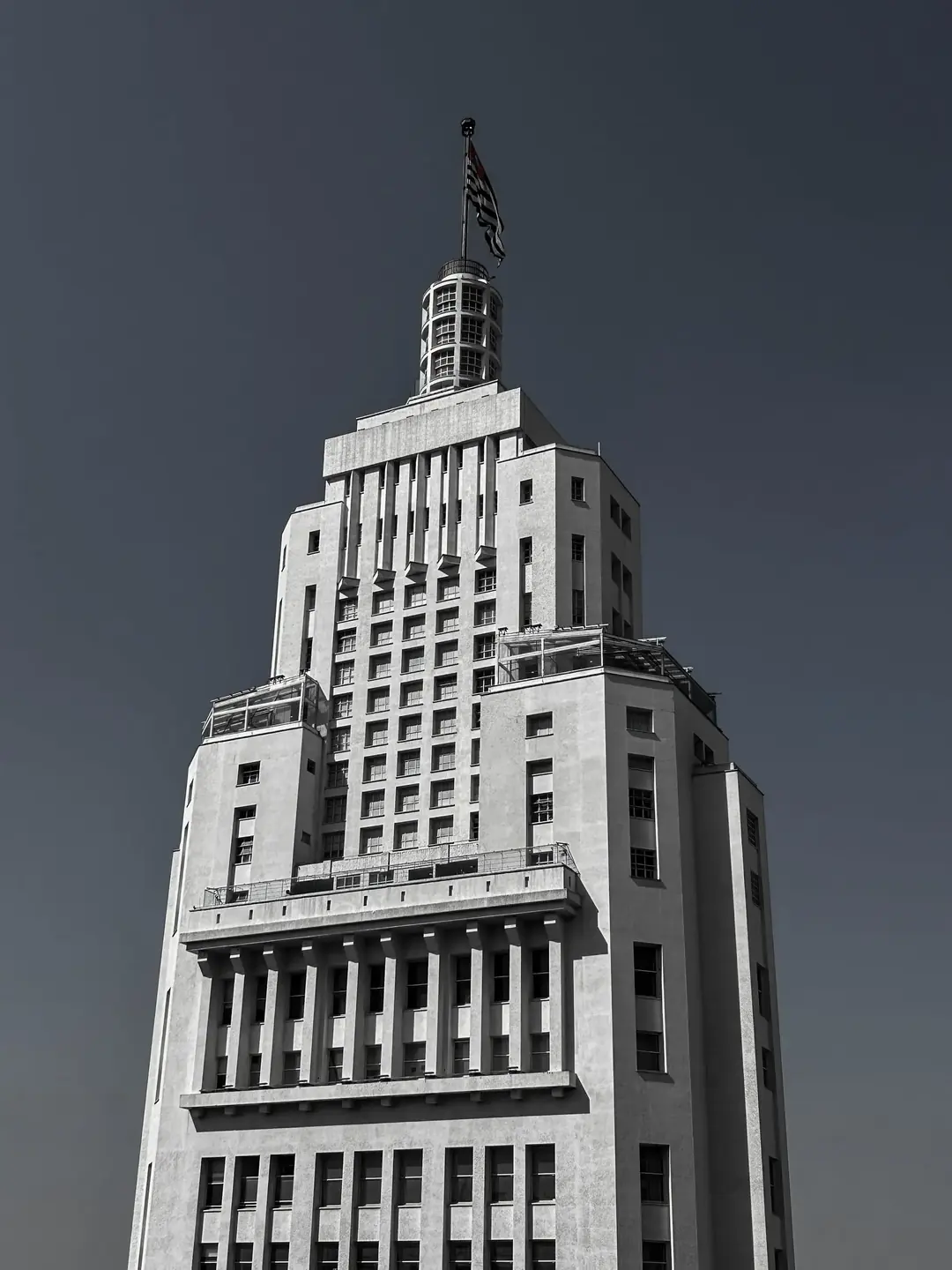 Topo do Edifício Altino Arantes, em São Paulo, em preto e branco, com a bandeira paulista ao vento contra o céu escuro